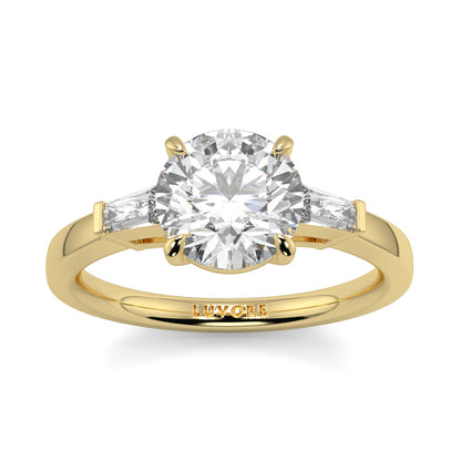 Gold ring with a large diamond and smaller diamonds on the shoulders, on a white background. yb_jewellers_3_stone_trilogy_engagement_ring_round_and_pear_shape_best_selling_design_trending_diamond_ring