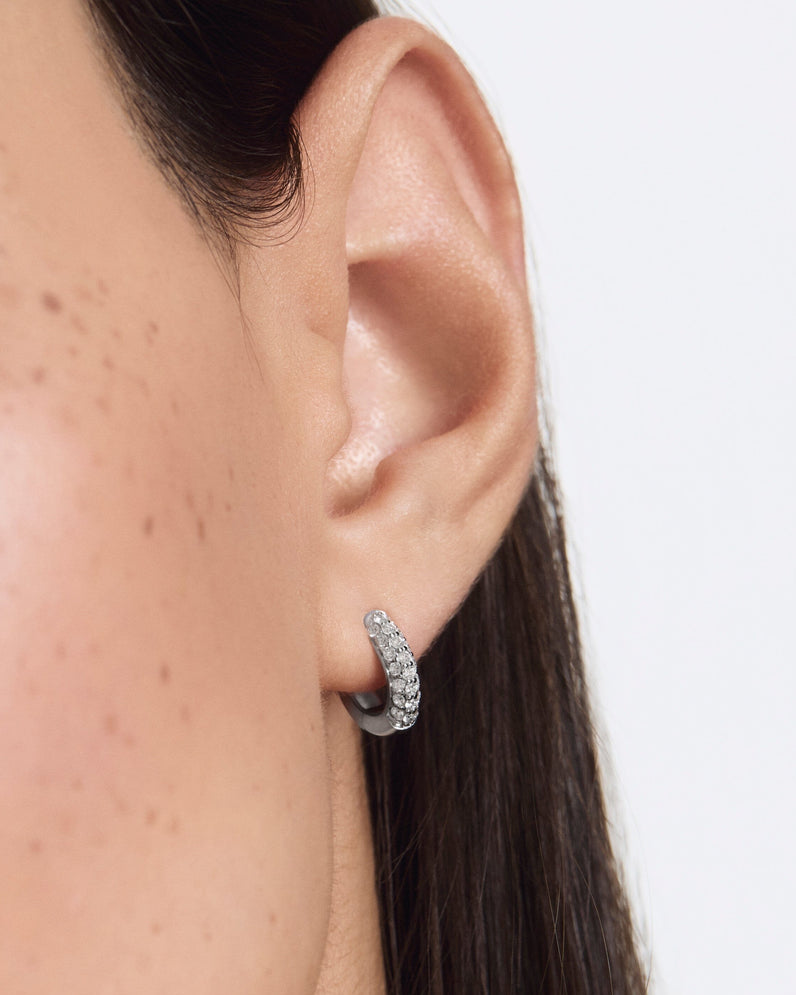 Close-up of an ear wearing a silver hoop earring on a plain background pair of huggiee earrings in 18k white gold platinum with micro diamond setting by yb jewellers 