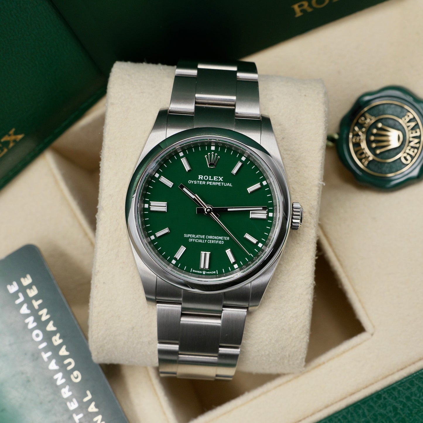 Rolex watch with green dial and silver bracelet on a beige cushion, surrounded by packaging elements.