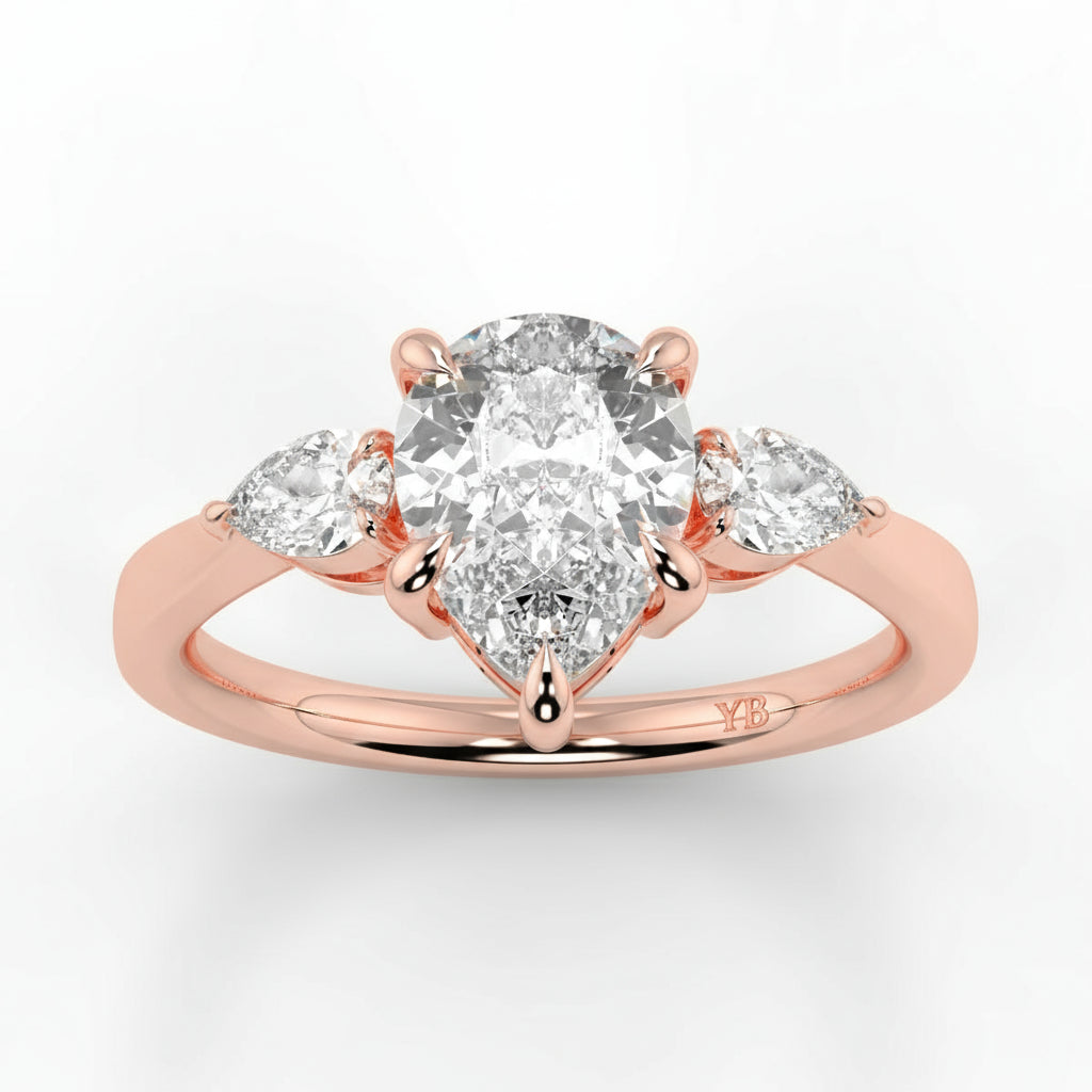 Rose gold ring with a large central diamond and two smaller diamonds on a light gray background _yb_jewellers_3_stone_trilogy_engagement_ring_round_and_pear_shape_best_selling_design_trending_diamond_ring