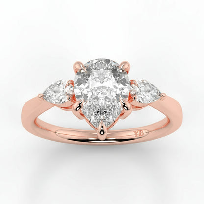 Rose gold ring with a large central diamond and two smaller diamonds on a light gray background _yb_jewellers_3_stone_trilogy_engagement_ring_round_and_pear_shape_best_selling_design_trending_diamond_ring