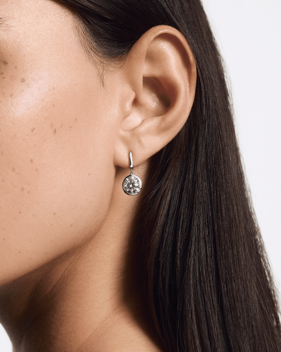 Close-up of an ear wearing a silver earring with a clear gemstone on a neutral background