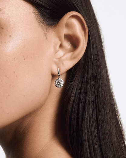 Close-up of an ear wearing a silver earring with a clear gemstone on a neutral background