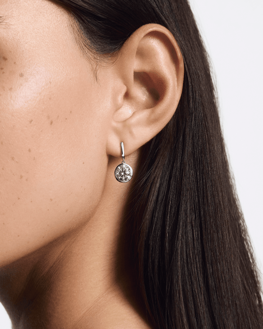 Close-up of an ear wearing a silver earring with a clear gemstone on a neutral background